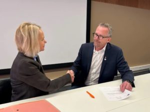 En mann og en kvinne som tar hverandre i hånden mens de sitter bak et bord. Mannen holder et papir i hånden.