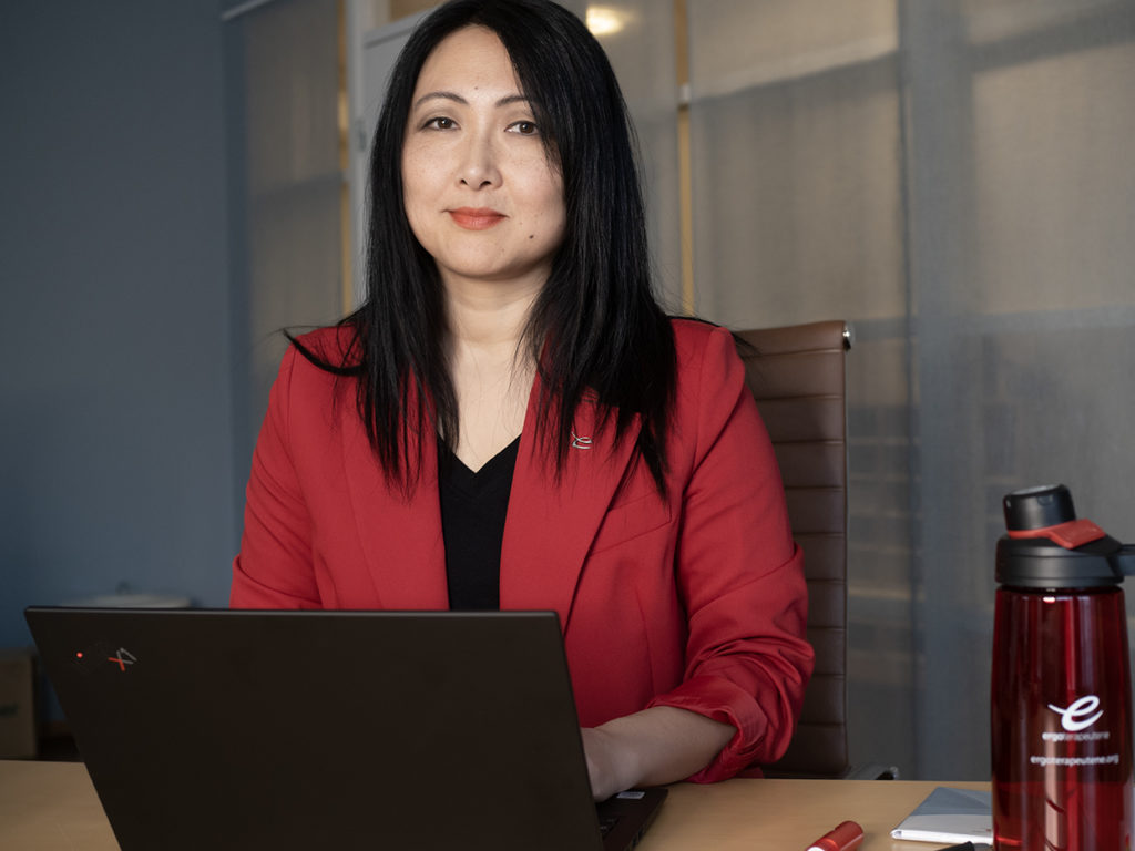 Kvinne med mørkt håpr og rød dressjakke. Sittende bak et bord med en laptop foran seg.